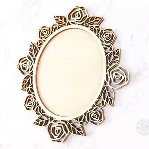 May include: A wooden oval frame with a floral rose design. The frame is made of light-colored wood with a laser-cut rose and leaf border. The frame is designed to hold a photo or artwork.