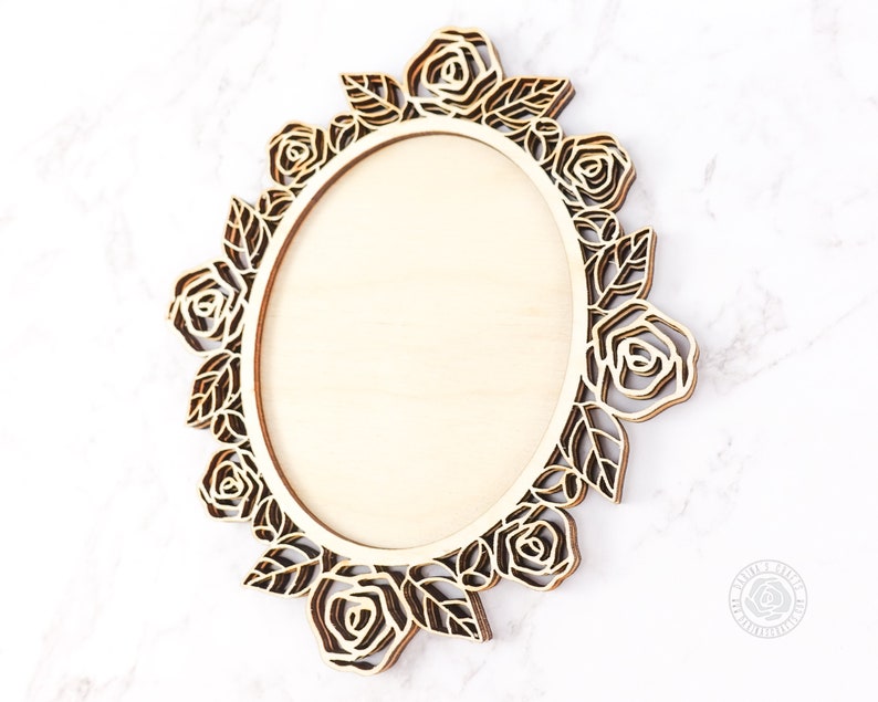 May include: A wooden oval frame with a floral rose design. The frame is made of light-colored wood with a laser-cut rose and leaf border. The frame is designed to hold a photo or artwork.