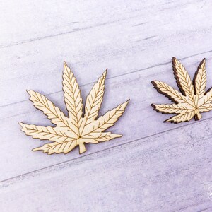 Puede incluir: Dos recortes de madera de hojas de marihuana, uno m&aacute;s grande que el otro, sobre un fondo de madera de grano claro gris.