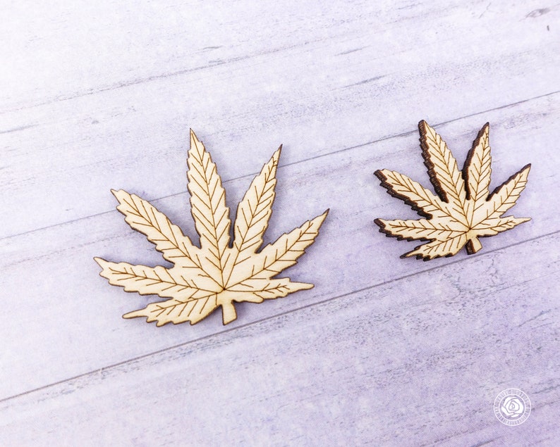 Puede incluir: Dos recortes de madera de hojas de marihuana, uno m&aacute;s grande que el otro, sobre un fondo de madera de grano claro gris.