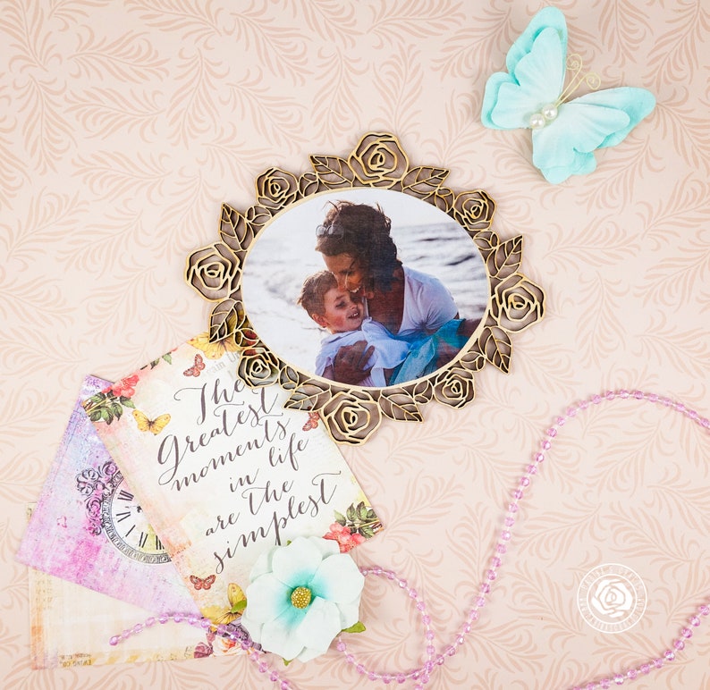 May include: A gold-toned decorative frame with a photo of a mother and child. The frame is surrounded by roses and leaves. The photo is set against a light blue background. The text "The greatest moments in life are the simplest" is written on a card with floral designs.