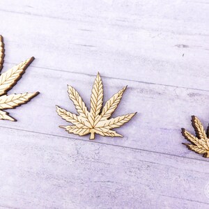 Puede incluir: Tres recortes de madera de hojas de marihuana de diferentes tama&ntilde;os. Las hojas son de color marr&oacute;n claro y tienen un dise&ntilde;o detallado.