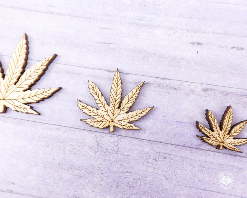 Puede incluir: Tres recortes de madera de hojas de marihuana de diferentes tama&ntilde;os. Las hojas son de color marr&oacute;n claro y tienen un dise&ntilde;o detallado.