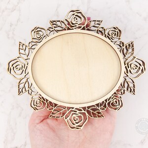 May include: A wooden oval frame with a floral rose design. The frame is made of light-colored wood and features laser-cut roses and leaves around the edge. The center is a smooth, blank oval, perfect for displaying a photo or artwork.