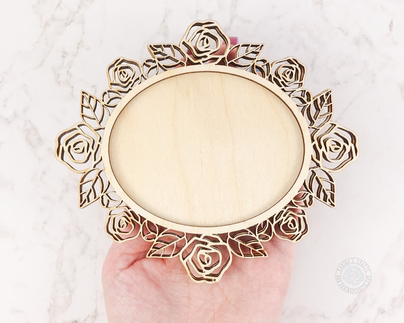 May include: A wooden oval frame with a floral rose design. The frame is made of light-colored wood and features laser-cut roses and leaves around the edge. The center is a smooth, blank oval, perfect for displaying a photo or artwork.