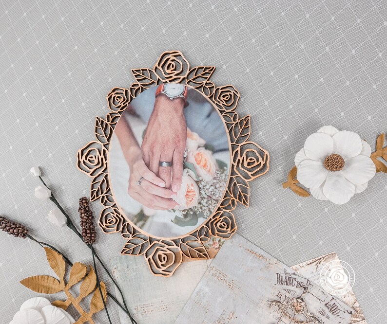 May include: A wooden picture frame with a rose design, featuring a photo of a couple's hands holding a bouquet of flowers. The frame is a light brown color and has a rustic look.