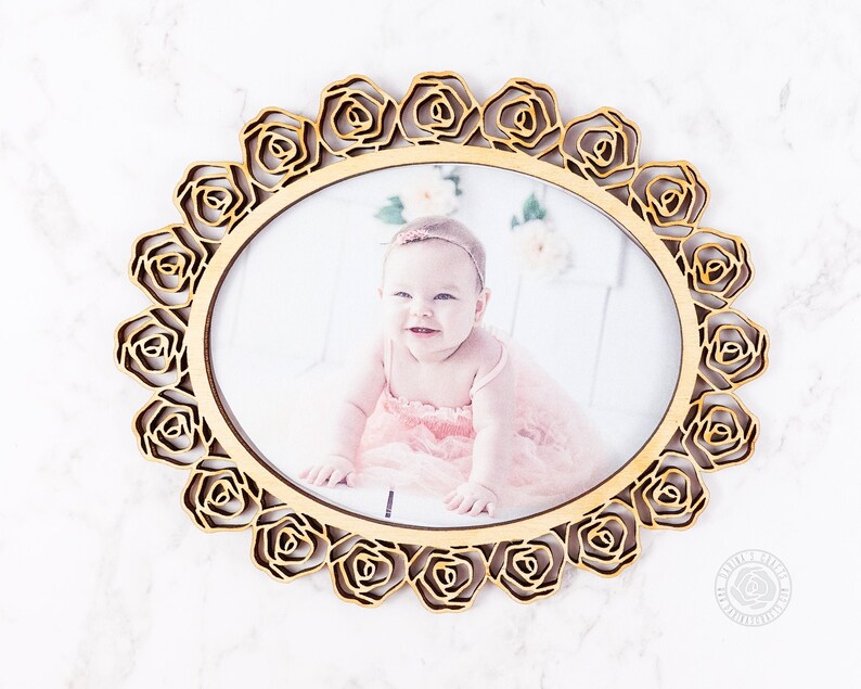 May include: A wooden photo frame with a rose design. The frame is light brown and has a circular shape. A photo of a baby girl is inside the frame.