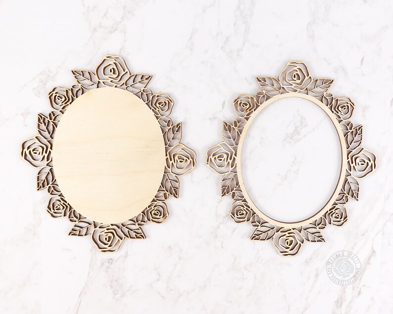 May include: Two wooden oval frames with intricate rose and leaf cutouts. One frame has a solid wooden center, while the other is open. The frames are light brown and sit on a white marble surface. The frames are decorative.
