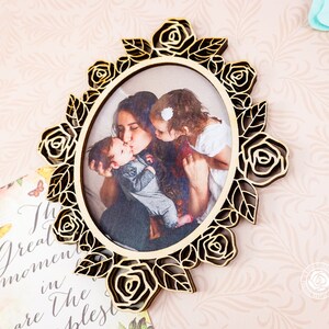 May include: Oval wooden picture frame with a laser-cut rose and leaf design. The frame holds a photograph of a mother kissing two children. The frame is made of light-colored wood.