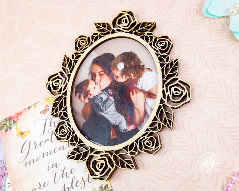 May include: Oval wooden picture frame with a laser-cut rose and leaf design. The frame holds a photograph of a mother kissing two children. The frame is made of light-colored wood.