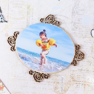 May include: A wooden photo frame with a floral design. The frame is oval shaped and has a photo of a child in a yellow life jacket standing in the ocean. The photo is surrounded by a decorative floral border.