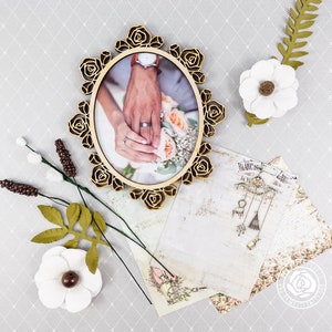 Puede incluir: Un marco ovalado de madera con un dise&ntilde;o floral, que presenta una foto de las manos de una pareja sosteniendo un ramo de flores. El marco est&aacute; rodeado de flores blancas y hojas verdes.