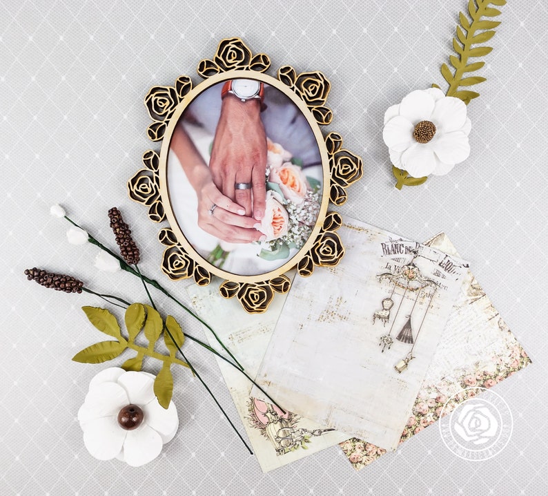Puede incluir: Un marco ovalado de madera con un dise&ntilde;o floral, que presenta una foto de las manos de una pareja sosteniendo un ramo de flores. El marco est&aacute; rodeado de flores blancas y hojas verdes.