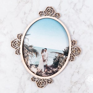 May include: A wooden oval photo frame with a cutout design of roses around the edge. The frame holds a photo of a couple standing on a beach.