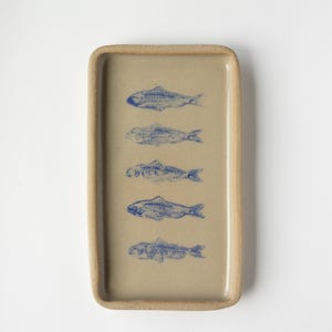May include: A rectangular ceramic tray with a beige base and a raised rim. The tray features five blue fish illustrations. The fish are arranged vertically, adding a decorative touch to the tray.