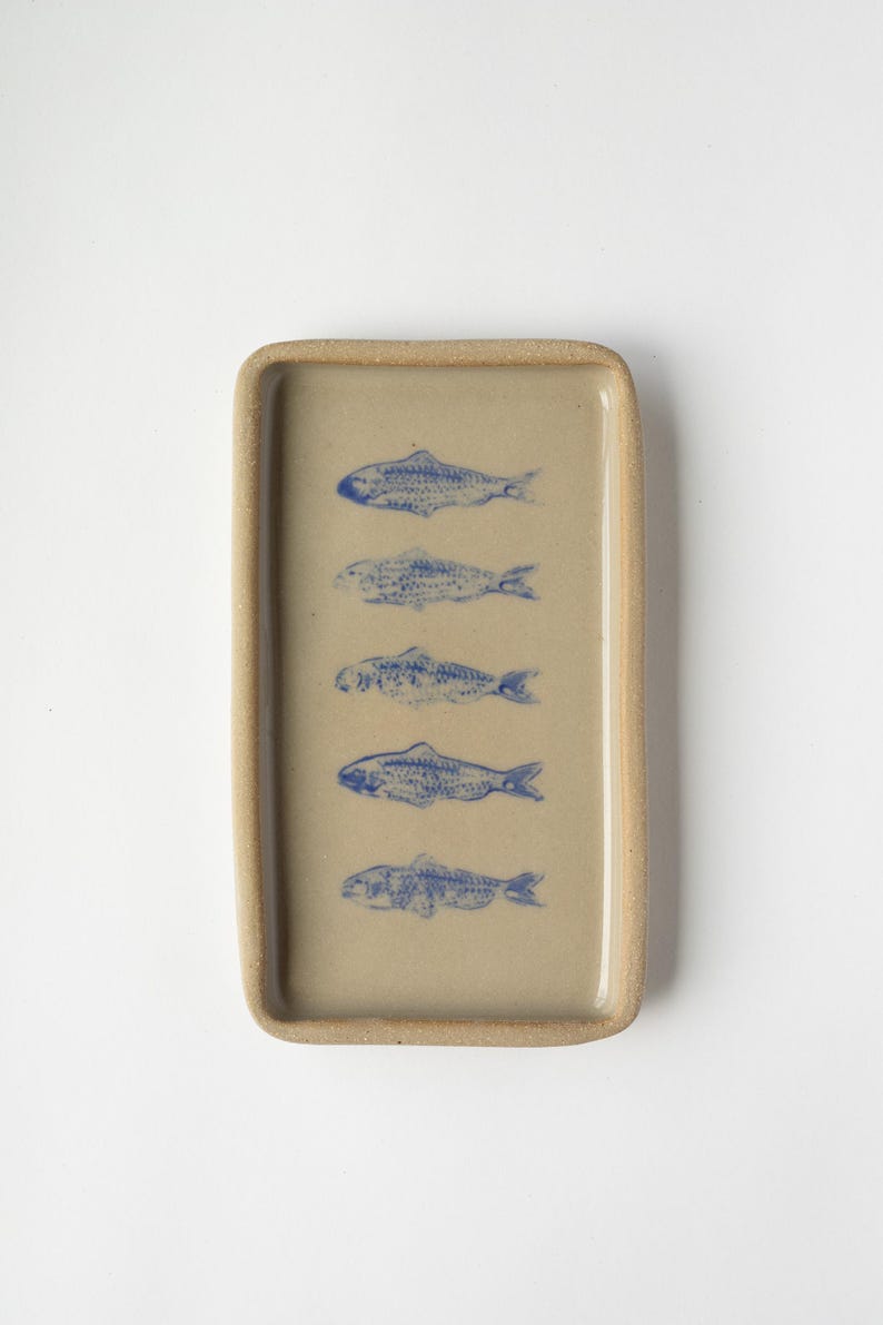 May include: A rectangular ceramic tray with a beige base and a raised rim. The tray features five blue fish illustrations. The fish are arranged vertically, adding a decorative touch to the tray.