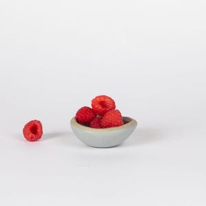 May include: A small, light blue ceramic bowl filled with red raspberries. The bowl is sitting on a white surface.