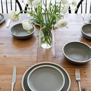 May include: A wooden dining table set for a meal. The table is set with gray plates, bowls, silverware, and a vase of white flowers. The plates have a white rim. The table is made of wood.