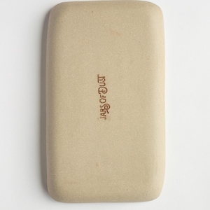 May include: A rectangular, beige ceramic tray with rounded corners. The text "JARS OF DUST" is printed in a dark brown color on the tray's surface. The tray has a smooth, matte finish.