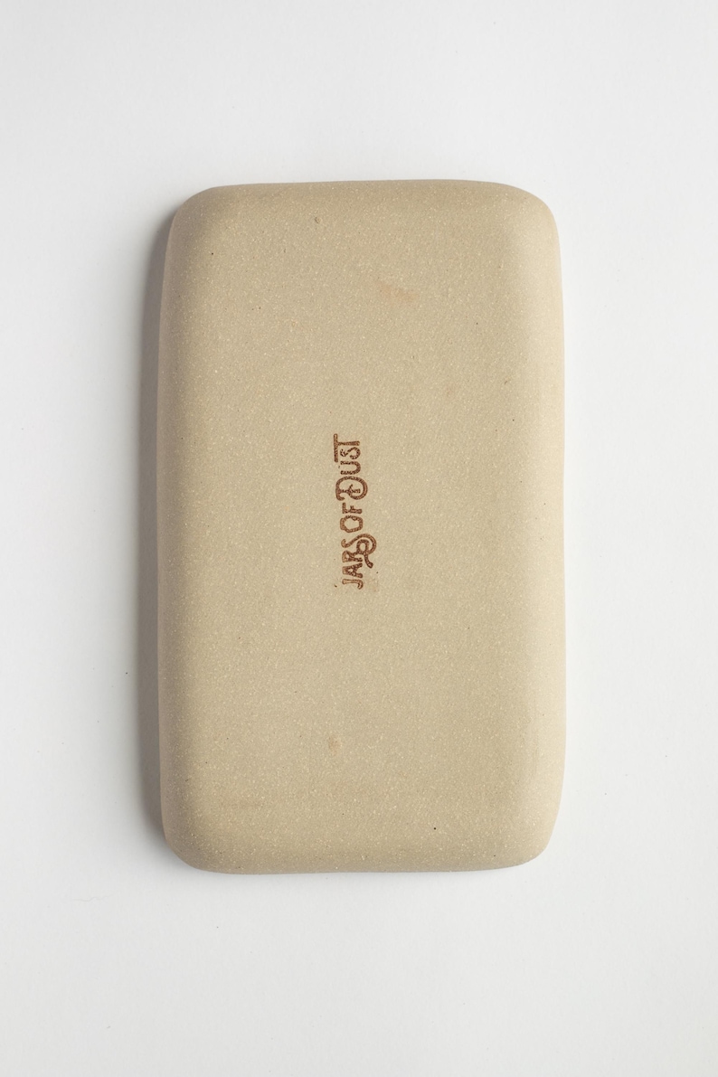 May include: A rectangular, beige ceramic tray with rounded corners. The text "JARS OF DUST" is printed in a dark brown color on the tray's surface. The tray has a smooth, matte finish.