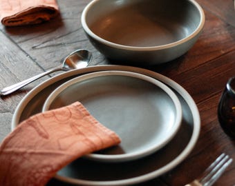 May include: A table setting featuring a gray bowl, plates, and a folded orange napkin. Silverware, including a spoon and fork, is arranged around the dishware. The items are set on a dark wooden table, creating a warm, inviting aesthetic.