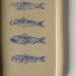 May include: Rectangular ceramic dish with a light beige surface. Five blue fish illustrations are evenly spaced across the surface. The dish has a slightly raised edge and a textured, light brown rim.