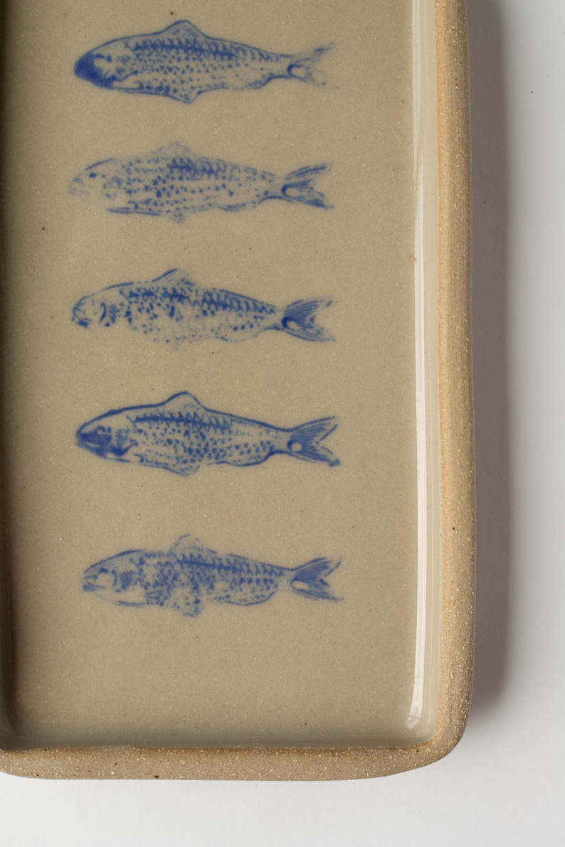 May include: Rectangular ceramic dish with a light beige surface. Five blue fish illustrations are evenly spaced across the surface. The dish has a slightly raised edge and a textured, light brown rim.