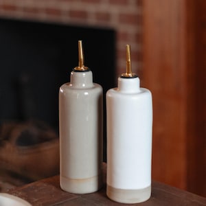 May include: Two ceramic oil and vinegar bottles with gold spouts. The bottle on the left is a light gray color and the bottle on the right is white. Both bottles have a brown glaze at the bottom.