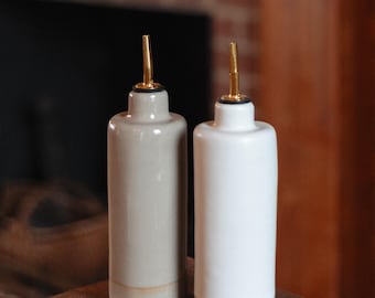 May include: Two ceramic oil and vinegar bottles with gold spouts. The bottle on the left is a light gray color and the bottle on the right is white. Both bottles have a brown glaze at the bottom.