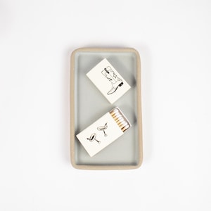 May include: Two white matchboxes with black illustrations on a light gray rectangular tray. One matchbox has a drawing of a cowboy boot and the other has a drawing of two martini glasses.