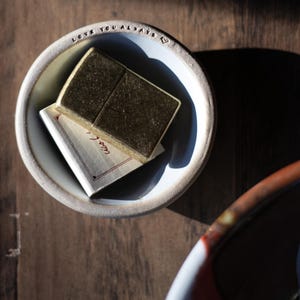 May include: A small, off-white ceramic bowl with the words "LOVE YOU ALWAYS" inscribed around the rim. Inside, a small, rectangular object is placed on top of a folded card. The object appears to be a solid, golden-brown color.