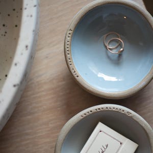 May include: Two ceramic bowls with light blue interiors and a light brown rim. One bowl contains two gold rings, and the other contains a small card with the text "ciao bella". The top bowl has the text "Will you marry me?" around the rim.