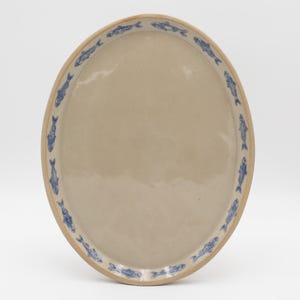 May include: An oval-shaped ceramic serving platter with a smooth, glossy beige center. The rim features a repeating pattern of small, detailed blue fish illustrations arranged in a circular border around the edge of the dish.