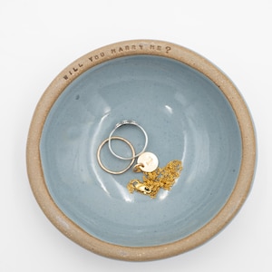 May include: A blue ceramic ring dish with the words "WILL YOU MARRY ME?" around the rim. The dish holds two rings and a gold necklace with a pendant.