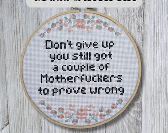 May include: A cross stitch kit with a wooden hoop. The white fabric features black text that reads "Don't give up you still got a couple of Motherfuckers to prove wrong." Pink and green floral accents surround the text. The text "Cross Stitch Kit" is at the top.