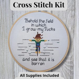 Peut inclure: Un kit de point de croix avec un tambour à broder en bois contenant un tissu blanc et un motif de l'épouvantail brodé. Le texte indique : Behold the field in which I grow my Fucks and see that it is barren. Le kit inclut toutes les fournitures.