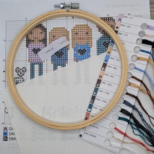 Peut inclure: Gros plan d'un projet de point de croix dans un cerceau de broderie en bois. Le projet repr&eacute;sente trois personnages avec un petit animal dans le coin inf&eacute;rieur gauche. Le cerceau est pos&eacute; sur une surface blanche avec un nuancier pour le fil &agrave; broder.