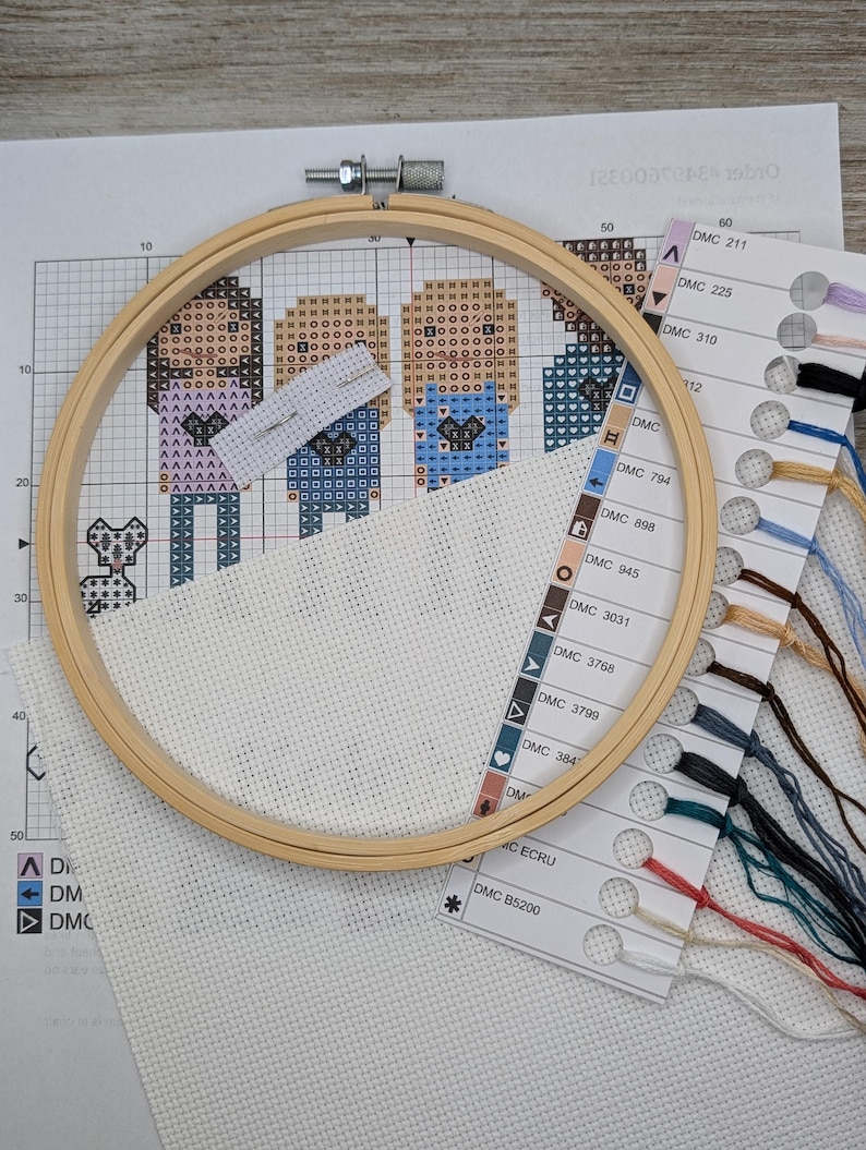 Peut inclure: Gros plan d'un projet de point de croix dans un cerceau de broderie en bois. Le projet repr&eacute;sente trois personnages avec un petit animal dans le coin inf&eacute;rieur gauche. Le cerceau est pos&eacute; sur une surface blanche avec un nuancier pour le fil &agrave; broder.