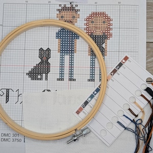 Peut inclure: Un motif de point de croix repr&eacute;sentant une famille de trois personnes et un chien. Le motif est sur une toile blanche quadrill&eacute;e avec un cerceau &agrave; broder en bois. Le motif comprend un homme, une femme et un chien. L'homme porte une chemise bleue et un pantalon bleu. La femme porte une chemise marron et un pantalon bleu. Le chien est noir. Le motif est accompagn&eacute; d'un tableau de couleurs avec les couleurs et les num&eacute;ros des fils.