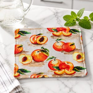 May include: A square fabric hot pad featuring a repeating pattern of whole and sliced peaches with green leaves on a light beige background. The design displays various orange and red peach illustrations with visible pits and textured skin.