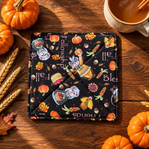 May include: A square fabric pot holder with a black background featuring a repeating pattern of mice, pumpkins, corn, and acorns. The text "Fall Blessings" and "Fellowship" is printed in a light color across the quilted surface.
