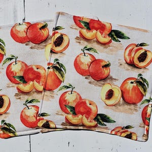May include: Two square fabric items with a repeating pattern of ripe peaches. The peaches are red and orange with green leaves, some whole and some sliced, on a light background. The items appear to be kitchen textiles.