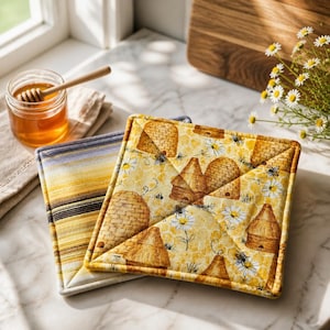 May include: Two square quilted pot holders. One has a yellow and white pattern with bees, daisies, and beehives. The other has yellow, black, and white horizontal stripes. A jar of honey and a honey dipper are in the background.