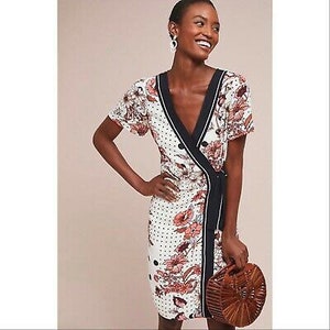 May include: A white wrap dress with a black and white polka dot pattern and a floral print. The dress has a V-neckline and short sleeves. It is tied at the waist with a black sash. The dress is paired with a brown woven bag.