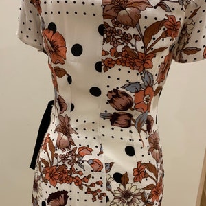 May include: A white dress with a floral and polka dot pattern. The dress has short sleeves and a V-neckline. The dress is cinched at the waist with a black ribbon.