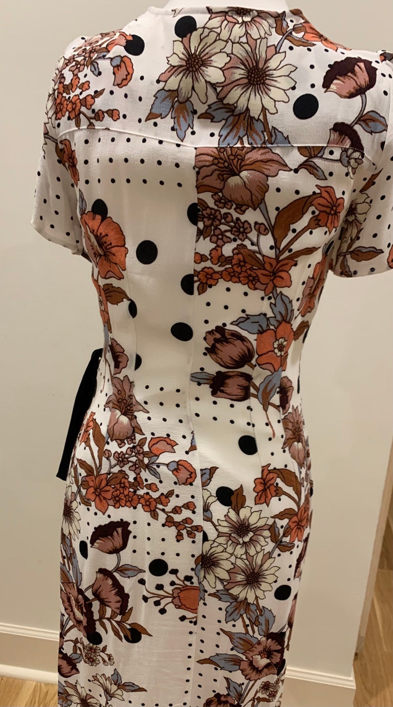 May include: A white dress with a floral and polka dot pattern. The dress has short sleeves and a V-neckline. The dress is cinched at the waist with a black ribbon.