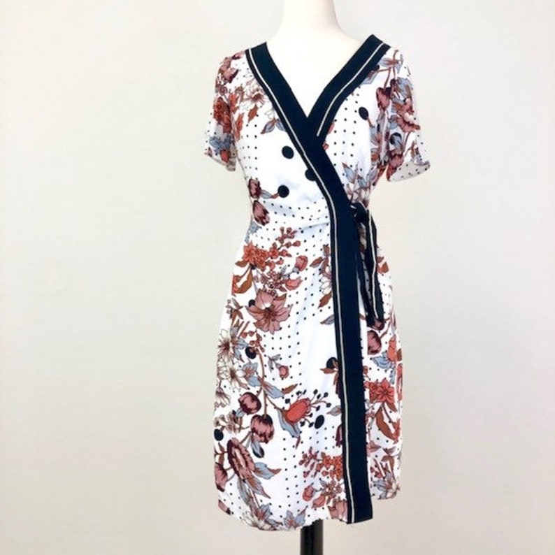 May include: A white wrap dress with a black and white polka dot pattern and a floral print. The dress has a black trim and a tie at the waist.