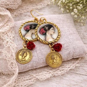 Può includere: Orecchini dorati con un ritratto di donna con un fiore rosso tra i capelli. Ogni orecchino presenta un ciondolo rotondo con un ritratto, una piccola rosa rossa e un ciondolo a forma di moneta d'oro. Gli orecchini sono esposti su un cuscino neutro.