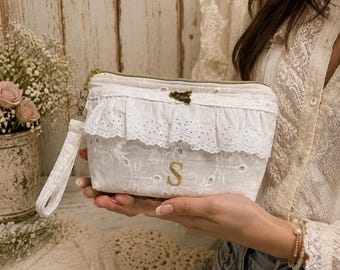 May include: A white zippered pouch featuring an embroidered floral pattern, a ruffled eyelet lace trim, and a gold-toned letter S monogram. The accessory includes a matching fabric wrist strap and a small gold-toned metal charm attached to the zipper.