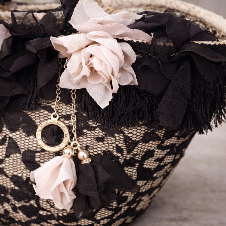 Pu&ograve; includere: Primo piano di una borsa decorativa con un motivo in pizzo nero e beige. La borsa &egrave; adornata con tessuto nero, frange e fiori di tessuto rosa chiaro. Una catena dorata con nappe rosa e nere pende dalla borsa.